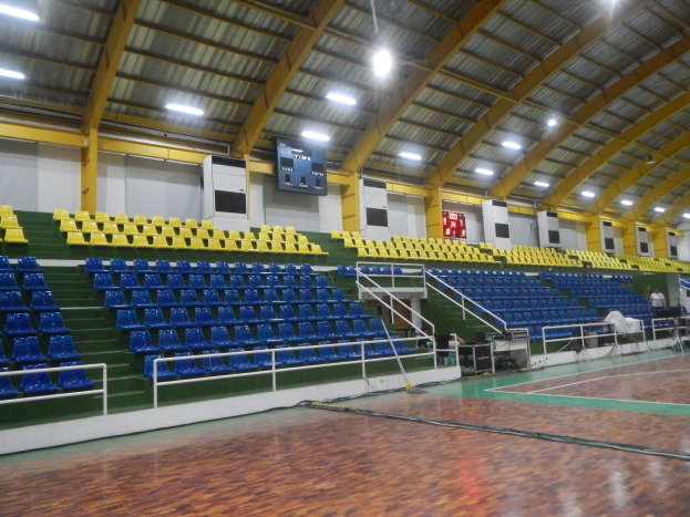 Große Sporthalle mit Holzboden, blauen und gelben Sitzplätzen, Geländern, Treppen mit Geländern, Wandbildschirmen, Deckenleuchten und einigen Personen.