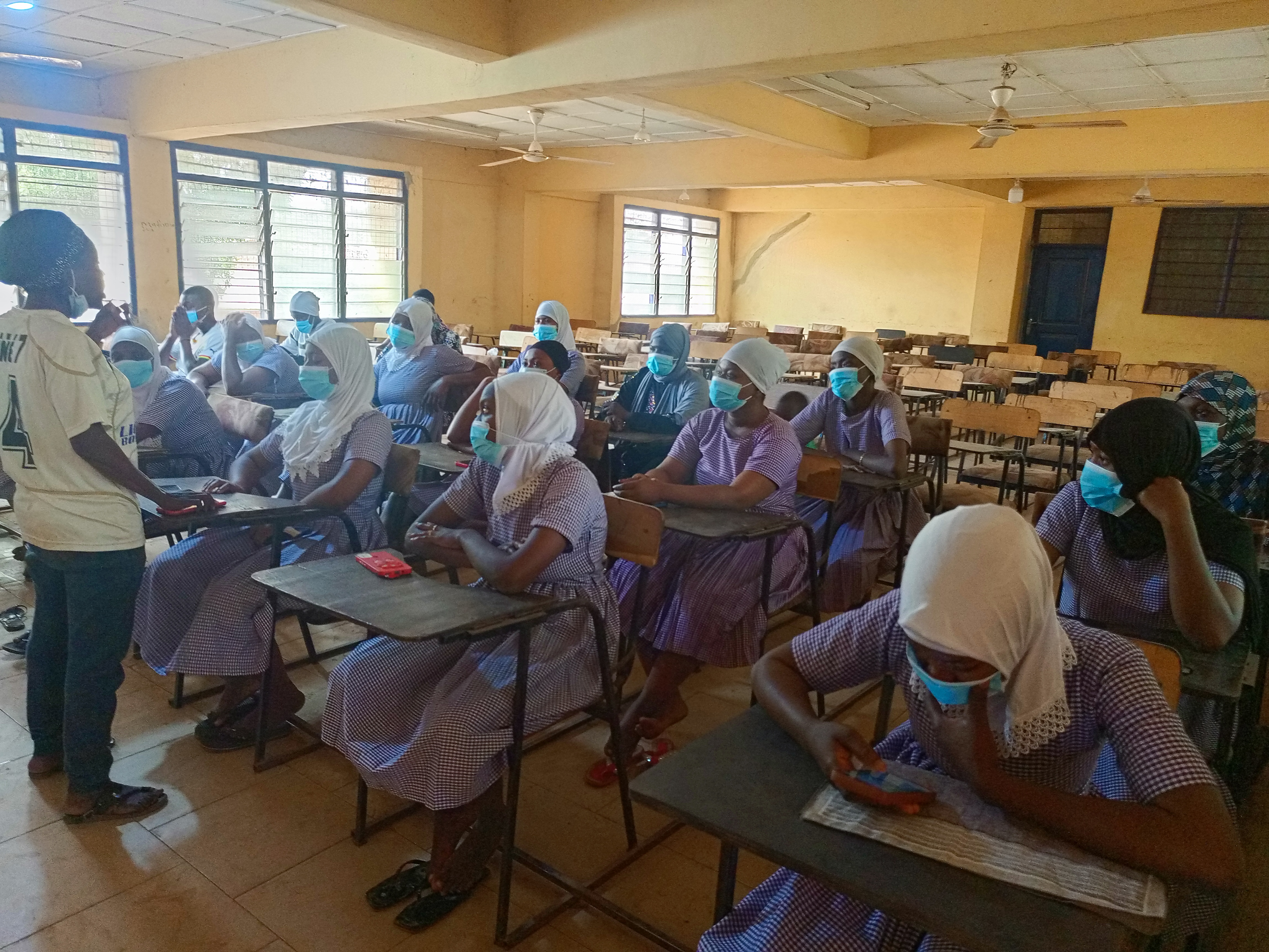 Eine Klasse in Nigeria mit Schülerinnen und Schülern, die Gesichtsmasken tragen und auf Bänken sitzen oder stehen, mit Büchern und Gegenständen auf den Tischen, Fenstern, einer Tür und Deckenleuchten und -ventilatoren im Hintergrund.