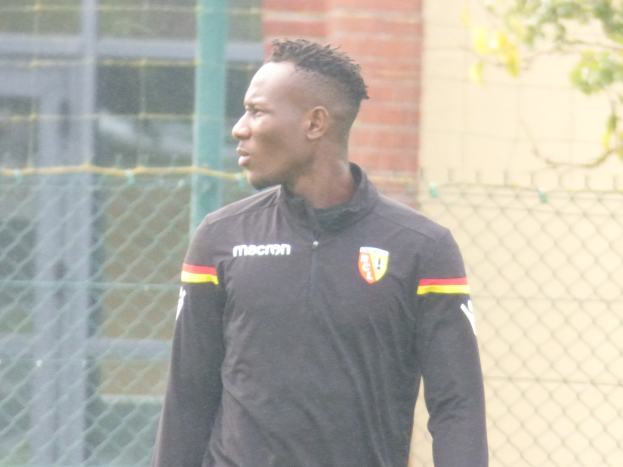 Ein Mann in einer schwarzen Jacke steht auf einem Fussballfeld, umgeben von einem Zaun und einem Baum, und sieht konzentriert während eines Trainingssitzungs.