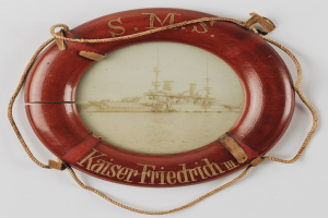 Rotes Rettungsflo├č mit 'USS Kaiser Friedrich III'-Text und einem Bild eines Schlachtschiffs, an einer wei├čen Oberfl├Ąche mit Seilen befestigt.