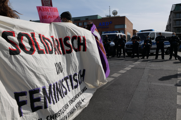 Eine Gruppe von Menschen marschiert auf einer Straße, hält eine 'Solidarität und Feminismus'-Schleife, mit geparkten Fahrzeugen, Gebäuden mit Fenstern, einer Schüsselantenne und einem klaren blauen Himmel im Hintergrund.