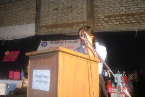 Ein Mädchen in einem weißen Hemd steht in der Nähe eines Rednerpults in einem Konferenzraum, rechts daneben ein Mikrofonständer, ein weißes Banner an einer Wand im Hintergrund und mehrere Taschen an einem Faden rechts hängend.