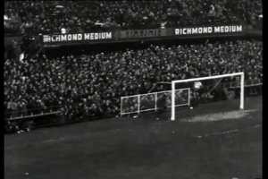 Ein Schwarz-Weiß-Foto eines Fußballspiels in einem Stadion, mit Zuschauern auf den Rängen und Spielern auf dem Feld, das ein Tor mit Netz und ein Anzeigebrett mit der Aufschrift "West Ham United v West Ham United - FA Cup Final" zeigt.