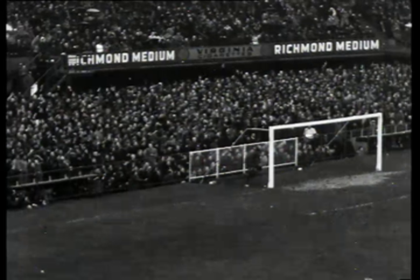 Ein Schwarz-Weiß-Foto eines Fußballspiels in einem Stadion, mit Zuschauern auf den Rängen und Spielern auf dem Feld, das ein Tor mit Netz und ein Anzeigebrett mit der Aufschrift "West Ham United v West Ham United - FA Cup Final" zeigt.