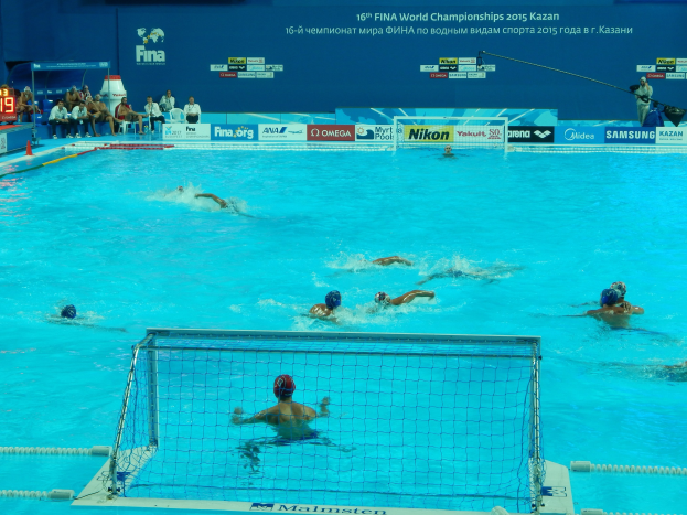 Gruppe von Athleten, die Wasserball in einem Pool während der 2015 FINA-Weltmeisterschaften in Kazan spielen, mit einem Netz, das den Pool teilt, und Zuschauern, die auf Stühlen im Hintergrund sitzen.
