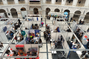 Große Besuchermenge auf einer internationalen Fachmesse mit Ständen, Bannern, Tischen, Stühlen und Luftballons in einem Raum mit architektonischen Bögen und Säulen.