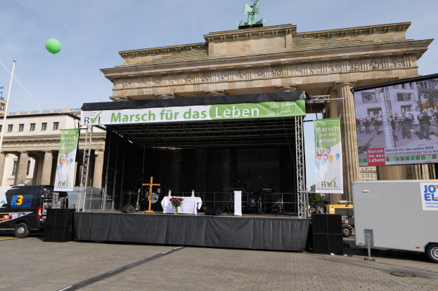 Bühne vor dem Brandenburger Tor in Berlin, Deutschland, mit einem Tisch mit einer Vase, einem Kreuzsymbol, Transparenten, Lautsprechern und anderen Gegenständen, sowie Fahrzeugen auf der Straße und Gebäuden im Hintergrund.