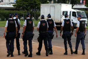 Eine Gruppe von Beamten in Uniform, einschließlich Mützen, Hemden, Schutzwesten, Hosen und Schuhen, geht auf einer Straße. Rechts daneben steht ein Lastwagen in der Nähe eines Zauns. Im Hintergrund sind Gebäude, Bäume, Pflanzen, Pfosten und Banner zu sehen. In der oberen rechten Ecke ist eine Brücke zu erkennen.