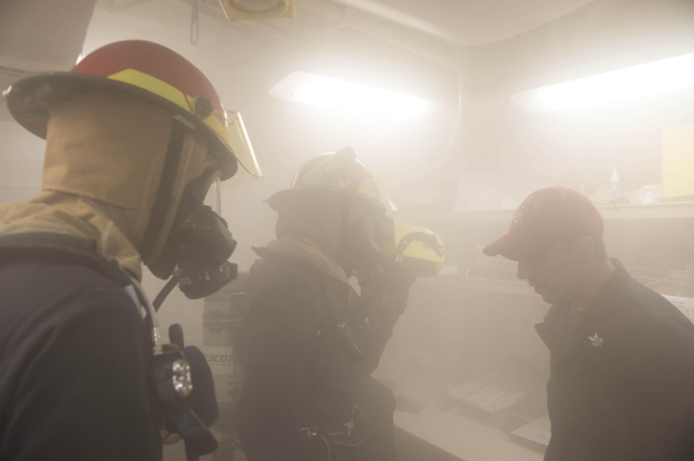 Feuerwehrleute in Helmen und Ausrüstung atmen Rauch während einer Übung aus, einer hält eine Kamera und Hintergrundbeleuchtung ist sichtbar.
