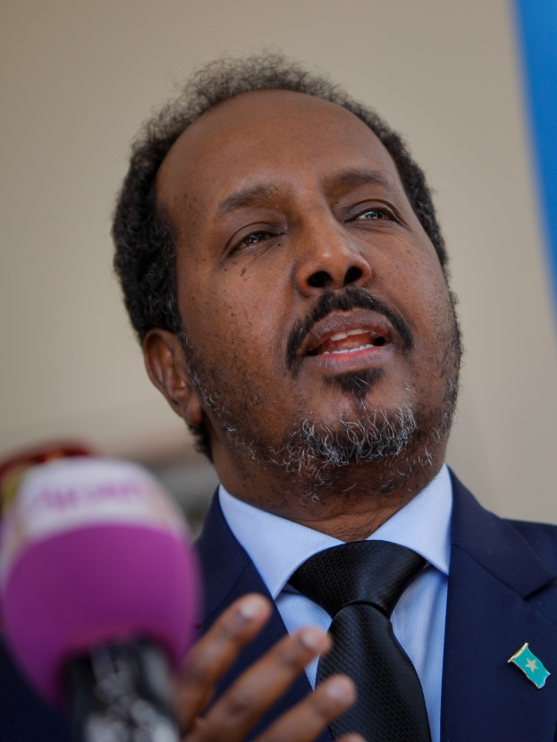 Ein Mann in Anzug und Krawatte, der in ein Mikrofon spricht, vor einem unscharfen Hintergrund, wahrscheinlich vor einem Publikum.