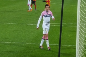 Ein Fussballspieler steht auf einem Feld neben einem Tor mit einem Netz auf der rechten Seite, während andere Spieler im Hintergrund sichtbar sind.