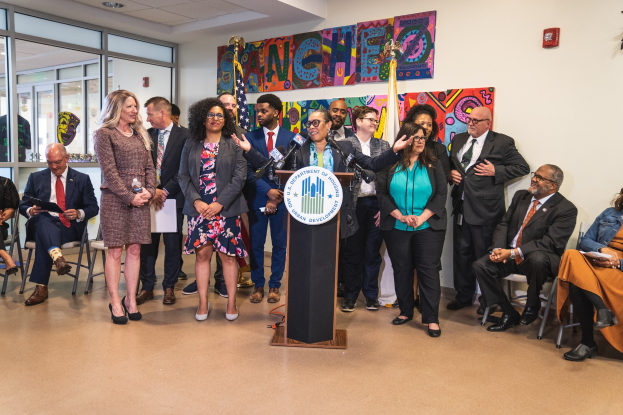 Gruppe von Menschen vor einem Podium mit einem Mikrofon und einer Tafel mit der Aufschrift "Sancho Stadtrat joins City Council", mit sitzenden und stehenden Teilnehmern, Flaggen, Feuerlöscher und Deckenleuchten.