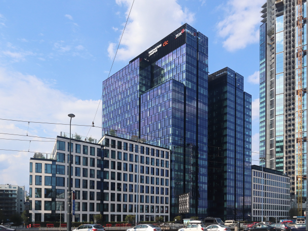 Großes Bürogebäude mit vielen Fenstern, identifiziert als Deutsche Bank Hauptsitz in Frankfurt, Deutschland, umgeben von städtischer Infrastruktur einschließlich Straßenlaternen, Fahrzeugen, Grünflächen und einem bewölkten Himmel.