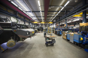 Ein großes Lagerinneres mit Maschinen, Fahrzeugen auf der linken Seite, Wagen mit Gegenständen, Holzboxen und anderen Gegenständen auf der rechten Seite und Lampen oben.