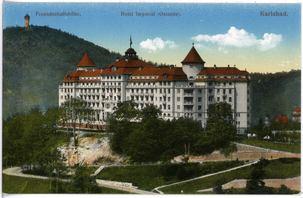 Alte Ansichtskarte des Hotels Imperial in Karlsbad, Deutschland, zeigt ein großes Gebäude mit Fenstern, umgeben von Bäumen, Pflanzen, einem Pfad, Hügeln und einem bewölkten Himmel, mit Text oben.