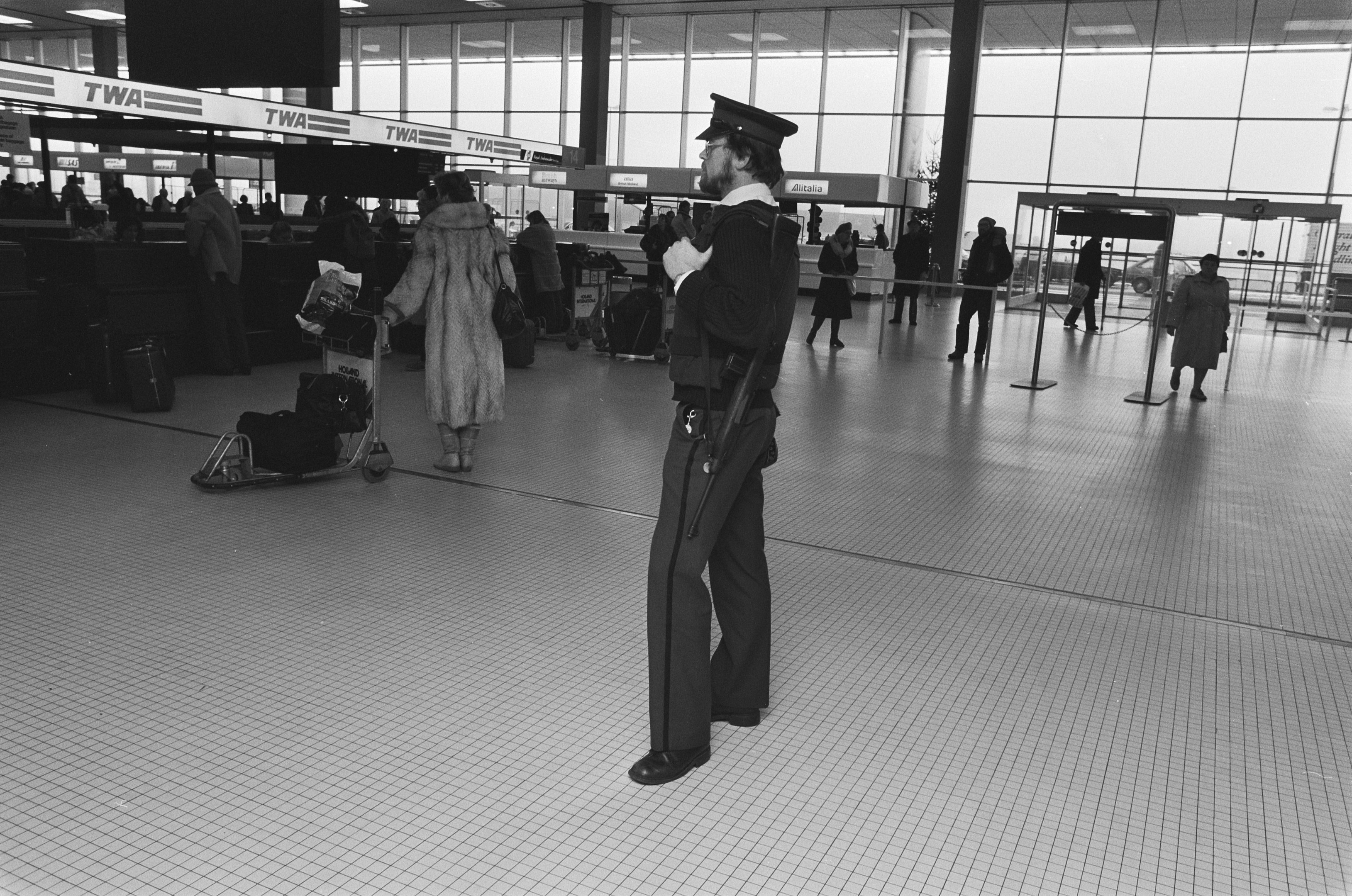 Ein Polizist in Uniform steht in einem Flughafen-Terminal, hält eine Waffe und ist von Menschen und Gepäck umgeben, mit Gepäckwagen, Schildern, Deckenleuchten, Pfosten und Glaswänden im Hintergrund.