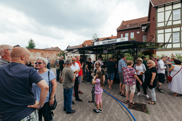 Menschen vor einem Gebäude mit Fenstern auf einem Bierfest, einige halten Gläser, mit Bäumen und einem bewölkten Himmel im Hintergrund.