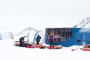 Drei Personen stehen auf einer schneebedeckten Landschaft mit Taschen drumherum, Zelten mit Skiern darauf und schneebedeckten Hügeln im Hintergrund bei klarem Himmel.