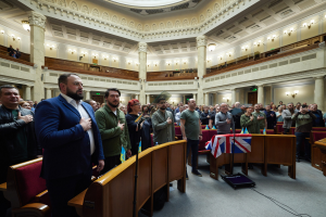 Eine Gruppe von Liberalen, die im House of Commons stehen, mit einer Menge, Stühlen, Tischen, Fahnen, einem Mikrofonständer, einem Gerät auf dem Boden, Säulen, Wandlampen und Deckenlampen im Hintergrund.
