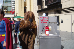 Eine Menschenmenge, darunter einige in Kostümen, versammelte sich vor Gebäuden mit Schildern, einer Box mit Aufschrift und einem Feuerlöscher.