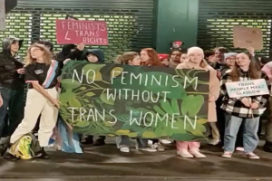 Eine Gruppe von Menschen, die eine Transparenz mit der Aufschrift "Kein Feminismus ohne Trans-Frauen" und Schilder in einem ├Âffentlichen Raum mit einer Wand und einem Rollladen im Hintergrund halten.
