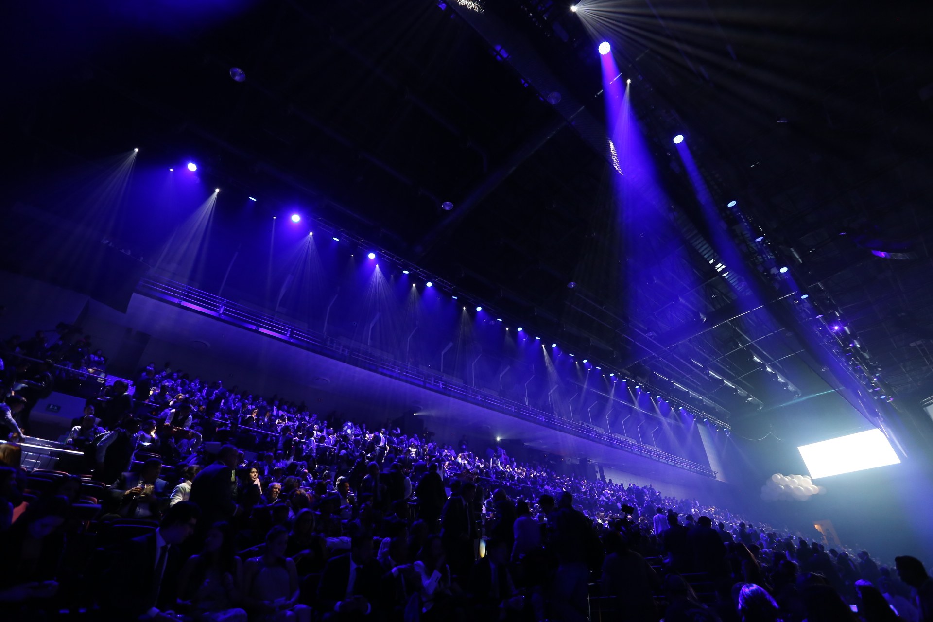 Eine große Menschenmenge sitzt in einem Konzertsaal gegenüber einer Bühne mit Deckenleuchten und einem Bildschirm auf der rechten Seite.