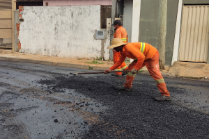 Zwei Männer in orangefarbenen Overalls und Hüten arbeiten an einer Straße, wobei einer ein Objekt hält, vor Gebäuden mit Fensterläden.