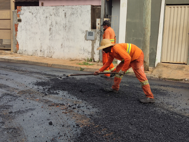 Zwei Männer in orangefarbenen Overalls und Hüten arbeiten an einer Straße, wobei einer ein Objekt hält, vor Gebäuden mit Fensterläden.