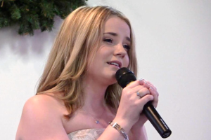 Eine Frau in einem weißen Kleid hält ein Mikrofon und singt vor einer Wand und Blättern im Hintergrund.