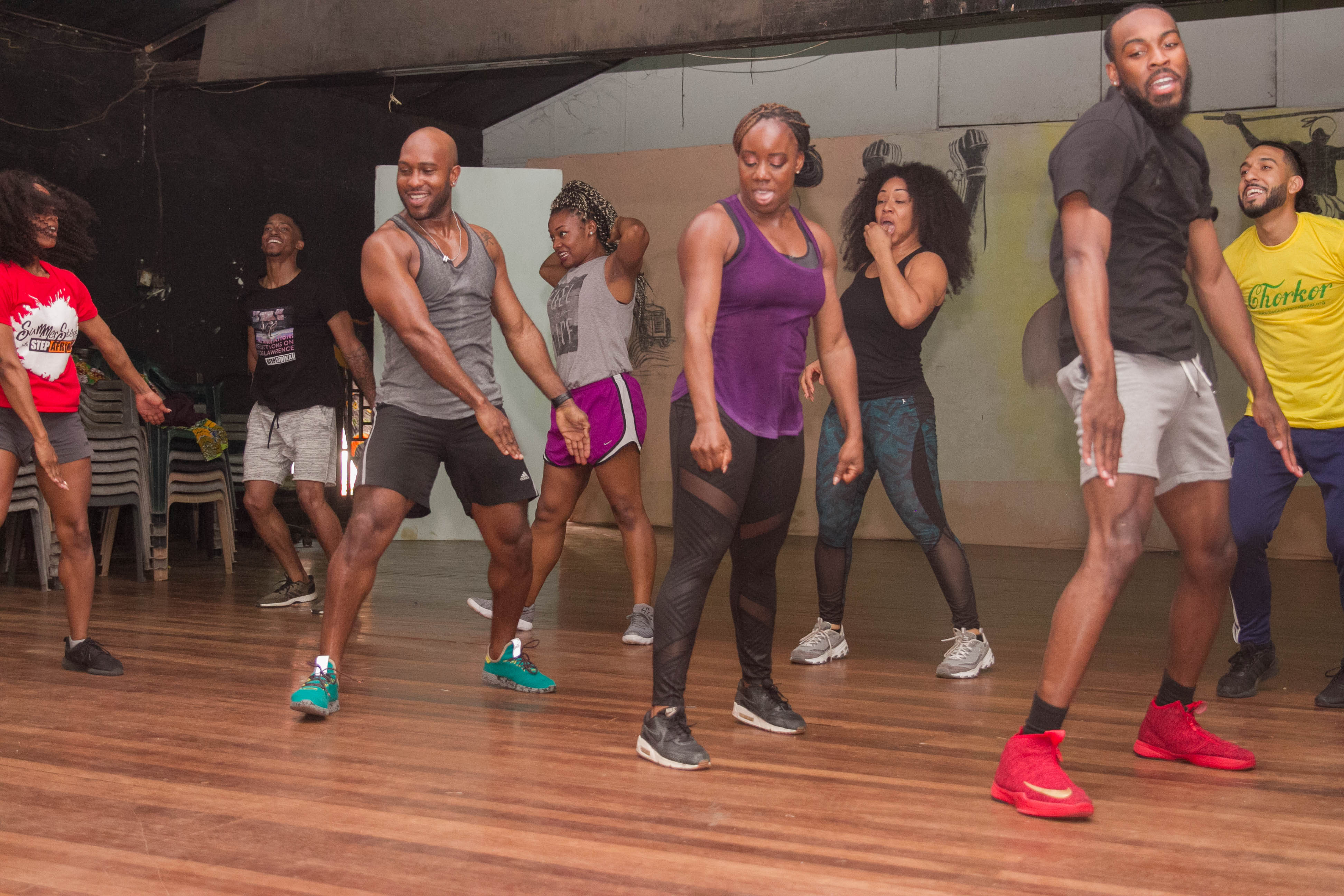 Eine Gruppe von Menschen, die in einem Tanzstudio tanzen, mit Turnschuhen bekleidet, einer bemalten Wand, Stühlen und anderen Gegenständen im Hintergrund, Deckenleuchten darüber und dem Boden darunter.