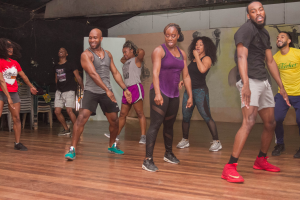 Eine Gruppe von Menschen, die in einem Tanzstudio tanzen, mit Turnschuhen bekleidet, einer bemalten Wand, Stühlen und anderen Gegenständen im Hintergrund, Deckenleuchten darüber und dem Boden darunter.