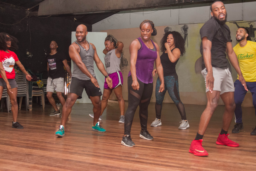 Eine Gruppe von Menschen, die in einem Tanzstudio tanzen, mit Turnschuhen bekleidet, einer bemalten Wand, Stühlen und anderen Gegenständen im Hintergrund, Deckenleuchten darüber und dem Boden darunter.