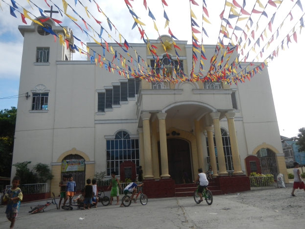 Eine Gruppe von Menschen, die Fahrräder vor einer weißen Kirche mit architektonischen Details, Pflanzen, einem Zaun, einer Fahne, Bäumen und einem bewölkten Himmel fahren, mit bunten Fähnchen an einem Seil oben.