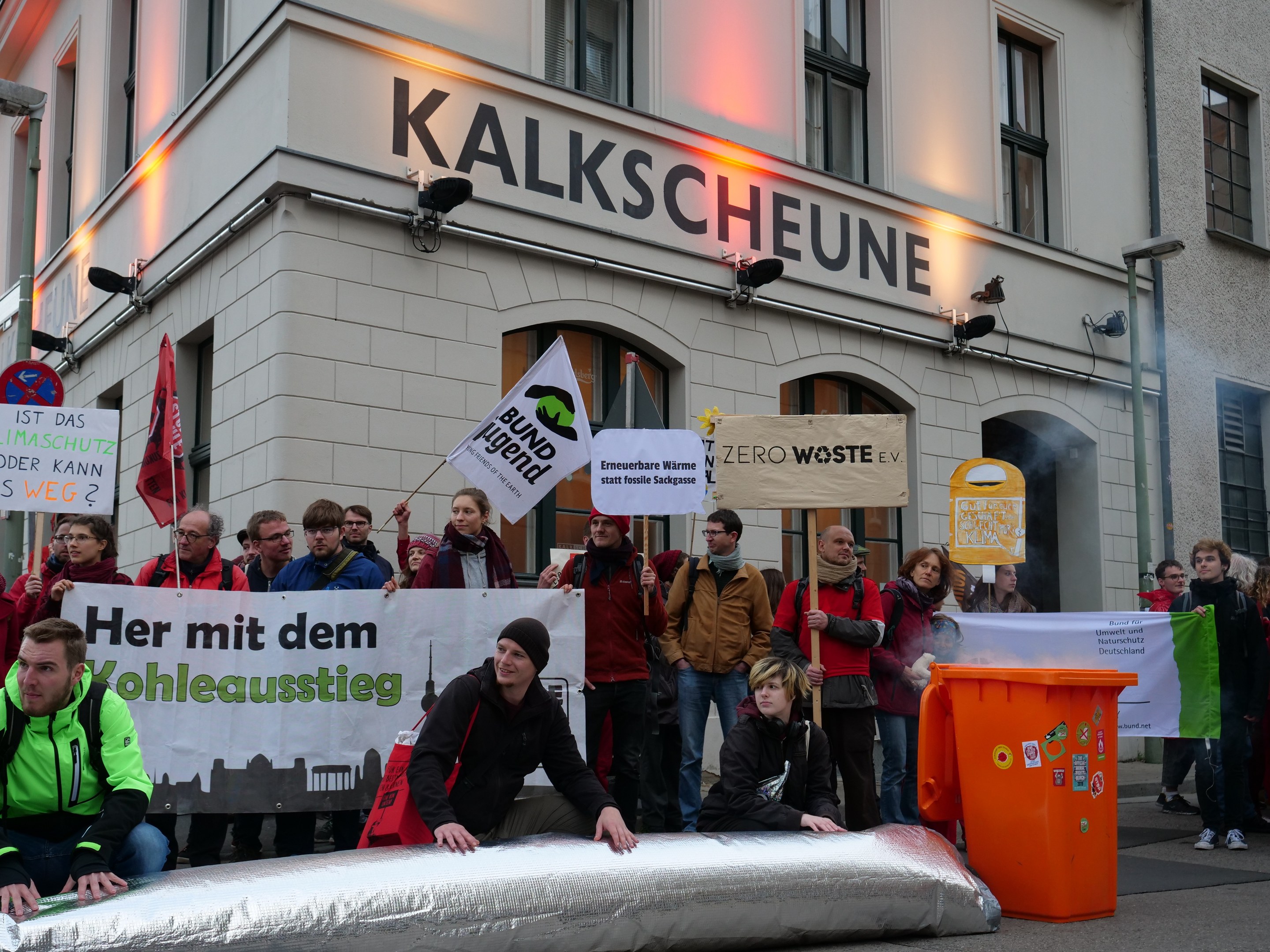 Eine Gruppe von Menschen mit Protestplakaten und Schildern vor einem Gebäude, mit zwei Personen auf einem Objekt im Vordergrund und einem Müllcontainer rechts.