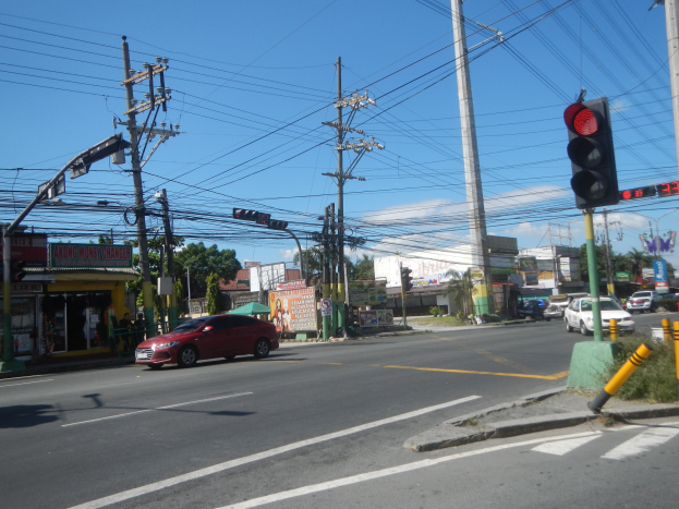 Ein rotes Auto fährt an einer Ampel auf einer Straße vorbei, die von Strommasten und -leitungen gesäumt ist, mit Gebäuden, Bäumen und einem klaren blauen Himmel im Hintergrund.