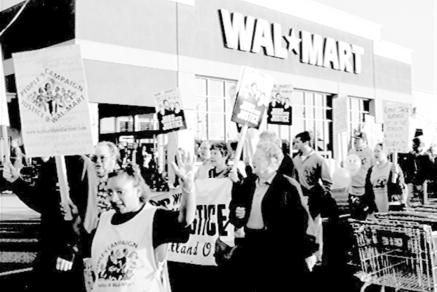 Schwarzes und weißes Bild von Menschen, die mit Plakaten protestieren, vor einem Einkaufszentrum im Hintergrund.