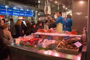 Marktszene mit Menschen, die gehen und Fische schneiden, Fische und Bretter unten und Geschäfte, Bretter, Stangen und Lichter im Hintergrund.