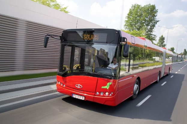 Roter Bus fährt auf einer Straße neben einem Gebäude mit einer Person darin, neben einer Wand, Bäumen, Laternenpfählen und einem bewölkten Himmel im Hintergrund.