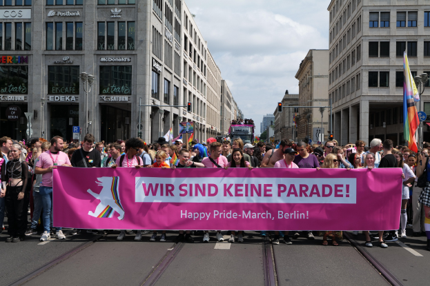 Eine Gruppe von Menschen marschiert auf einer Straße in Berlin, Deutschland, mit einer pinken Fahne, auf der "Happy Pride March" steht, während Gebäude, Laternenpfähle und Verkehrszeichen die Straße säumen und der Himmel bewölkt ist.