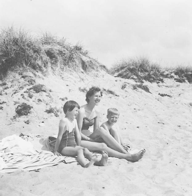 Schwarz-weißes Foto einer Frau in einem Bikini und zwei Kindern in Badebekleidung, die auf einer Decke auf einem sandigen Strand mit Gras und Himmel im Hintergrund sitzen.