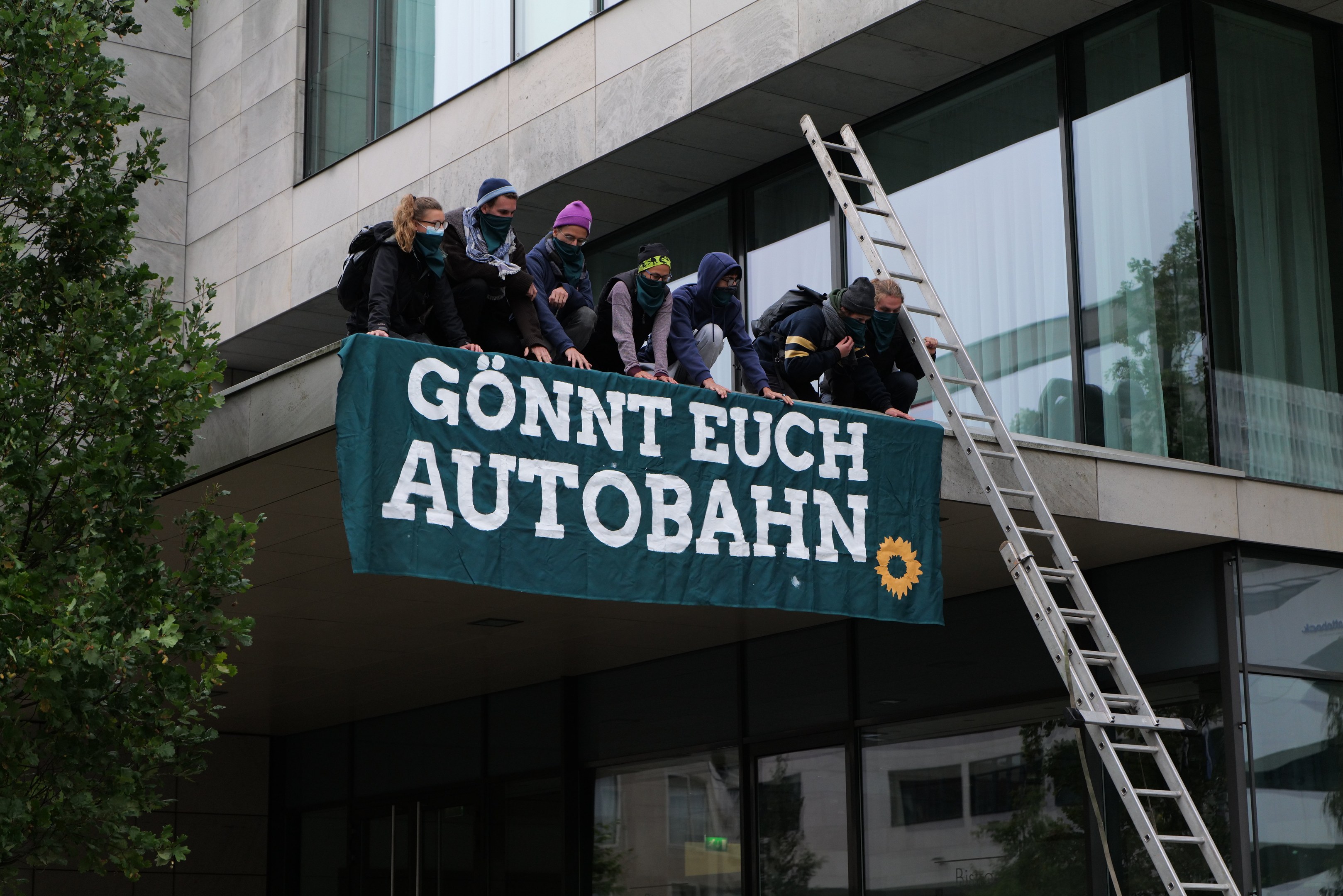 Eine Gruppe von Menschen auf einem Gebäude, die eine Fahne halten, mit einem Baum links und Glasfenstern im Hintergrund, wahrscheinlich protestieren sie für die deutsche Regierung.