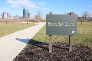 Ein verwittertes Schild am Boden in der Nähe einer grünen Fläche trägt die Aufschrift "Bocce Ball Courts in Indianapolis, Indiana" mit Bäumen, einem Zaun, Gebäuden und einem bewölkten Himmel im Hintergrund.