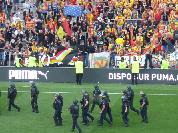 Eine Gruppe von Polizisten in Helmen und mit Waffen in der Hand geht auf einem Fussballfeld, im Hintergrund halten Menschen Fahnen und Transparente.