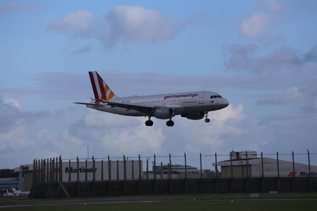 Germanwings Airbus A320-200 beim Start am Frankfurter Flughafen, mit Gras darunter, einem Zaun im Vordergrund, Gebäuden und Bäumen im Hintergrund und einer bewölkten Himmel über ihnen.