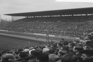 Schwarzes und weißes Foto eines vollen Stadions mit Zuschauern bei einem Fußballspiel, mit Bannern, Pfählen, einem Schuppen, Bäumen, einem Turm und einem bewölkten Himmel.