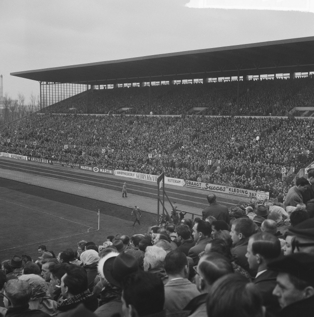 Schwarzes und weißes Foto eines vollen Stadions mit Zuschauern bei einem Fußballspiel, mit Bannern, Pfählen, einem Schuppen, Bäumen, einem Turm und einem bewölkten Himmel.