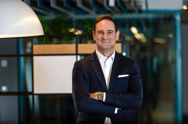 Ein Mann in einem weißen Hemd, schwarzer Jacke und Uhr steht lachend vor einer Glaswand, mit einem weißen Objekt links und einem unscharfen Hintergrund.