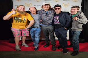 Fünf Männer in Anzügen auf einem roten Teppich, die lächeln und für ein Foto posieren, mit einem weißen "Speakeasy Awards 2019"-Banner und einem schwarzen Vorhang im Hintergrund.