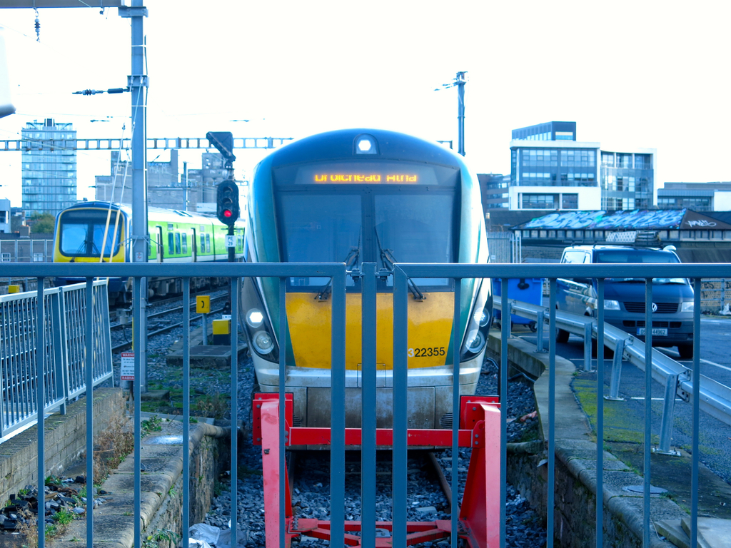 Zwei Züge auf den Schienen mit einem Zaun im Vordergrund, Pfosten mit einem, der ein Signallicht hat, ein Auto auf der Straße rechts, Gebäude im Hintergrund und Himmel darüber.