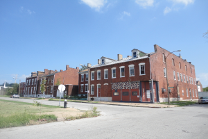 Ein Gebäude mit Pfählen und Straßenlaternen, umgeben von Bäumen und Fahrzeugen auf beiden Seiten, mit einem Gehweg, Gras und einem Feuerlöscher unten und Himmel oben.